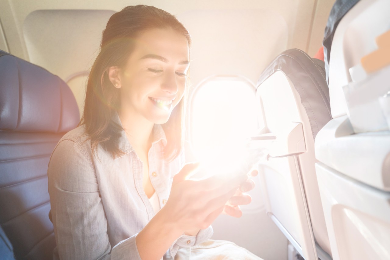 Cheerful young woman uses smart phone while on a commercial flight. The sun is streaming through the window next to her.